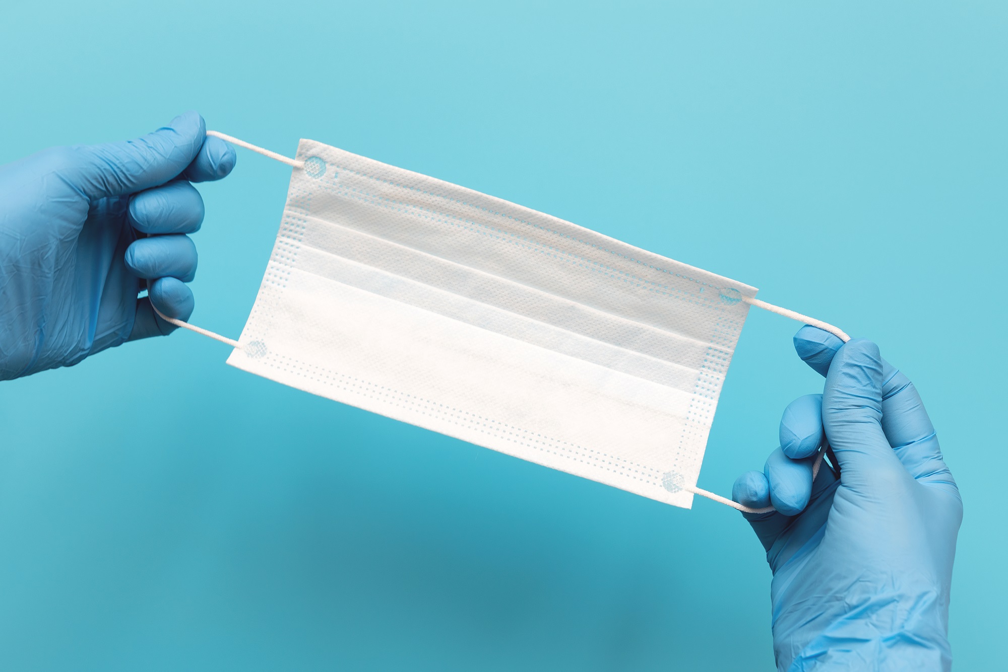 Hands of medic wearing blue latex gloves holding surgical ear-loop mask on blue background. Protection concept