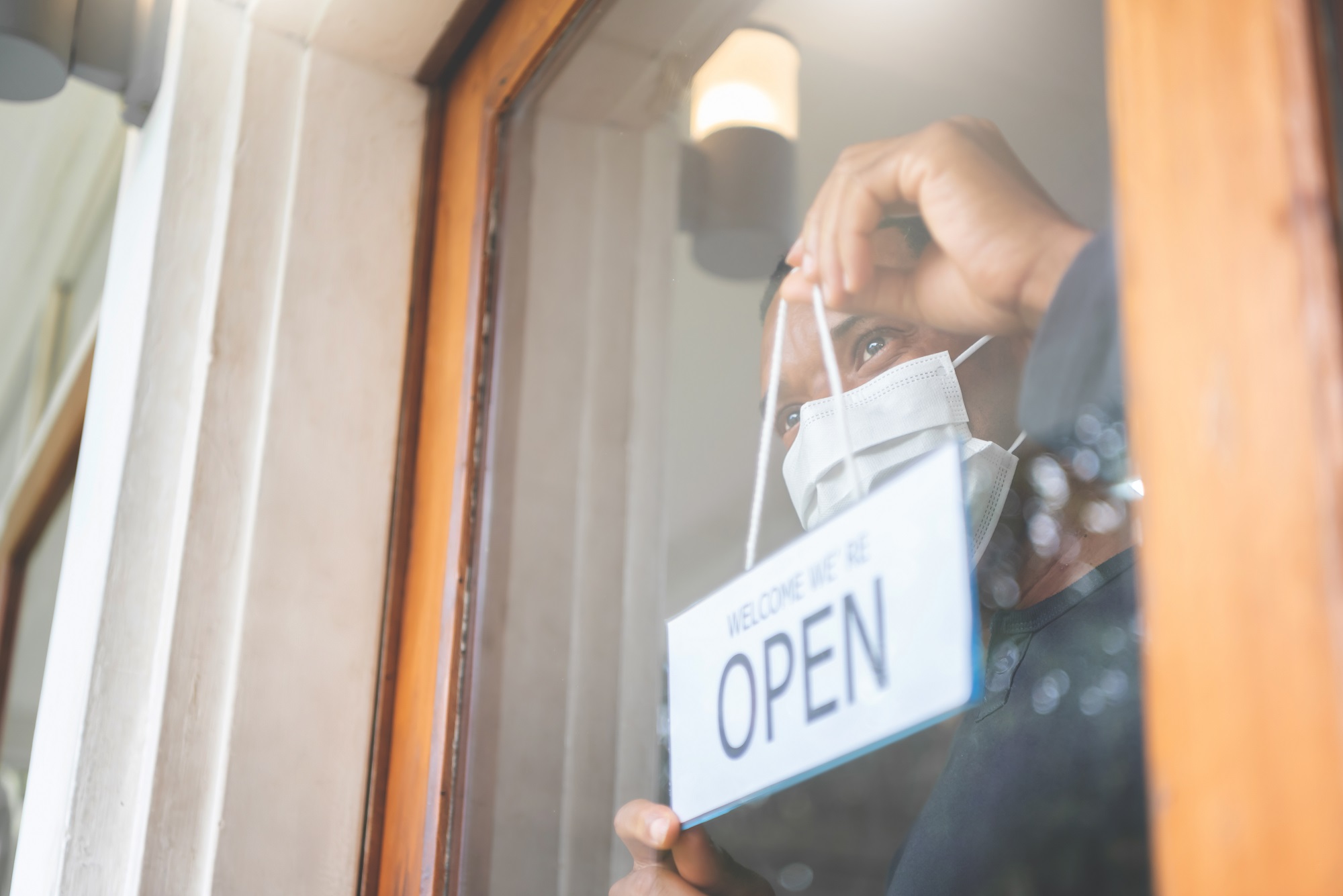 Black male holding open sign and looking out at the glass of the door. African American cafe man owner with face mask reopening after pandemic virus. Startup small business after Covid-19 quarantine.