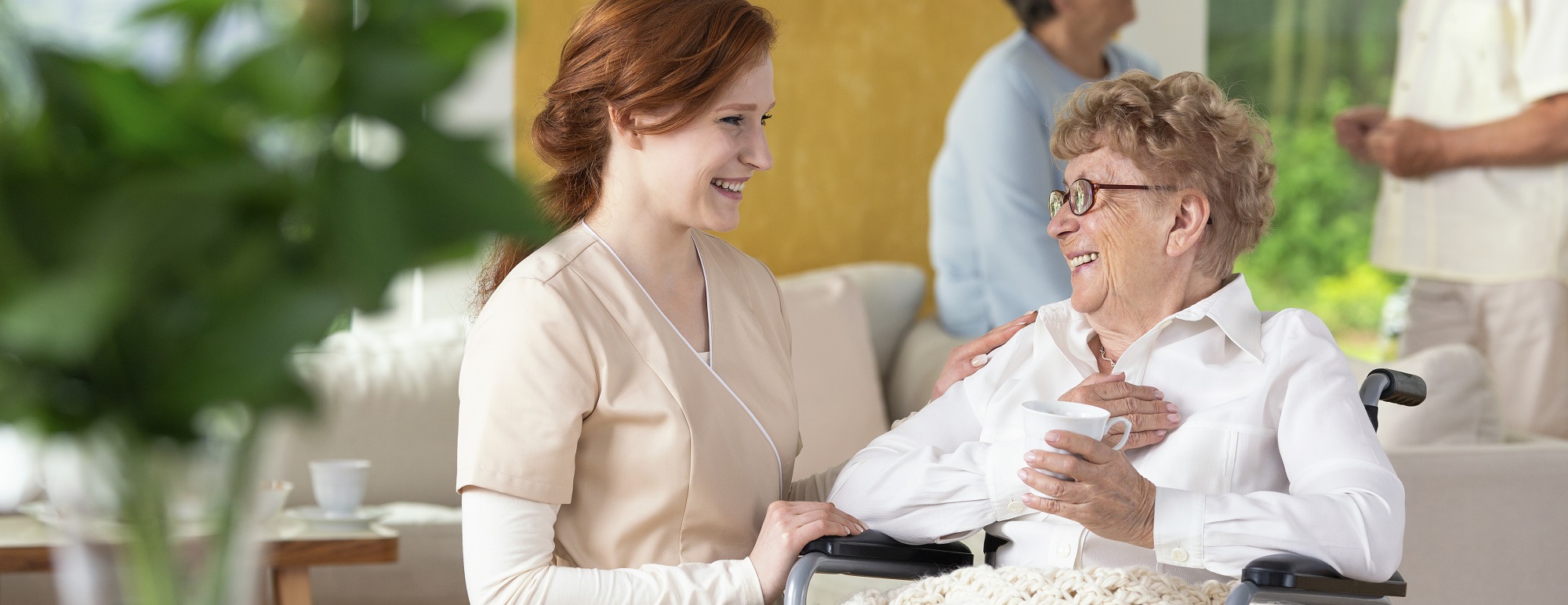 Young caretaker making a geriatric pensioner in a wheelchair laugh during leisure time in a common room of a luxury rehabilitation center. Blurred surrounding. Panorama.