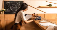 Side view of concentrated café/ small business owner with papers using payment terminal near calculator on table.