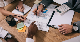 Engaging image captures a collaborative business meeting, showcasing diverse hands pointing at data charts and graphs. The warm wooden table contrasts with colorful sticky notes and notebooks, while a calculator adds practicality to the setting. The mood exudes professionalism and teamwork, ideal for corporate presentations or marketing materials. DEPOST PHOTOS