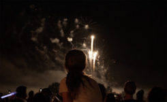 A little girl sits on someone's shoulders watching fireworks at Bell Works from above the crowd.