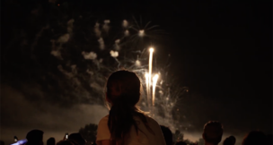 A little girl sits on someone's shoulders watching fireworks at Bell Works from above the crowd.