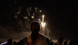 A little girl sits on someone's shoulders watching fireworks at Bell Works from above the crowd.